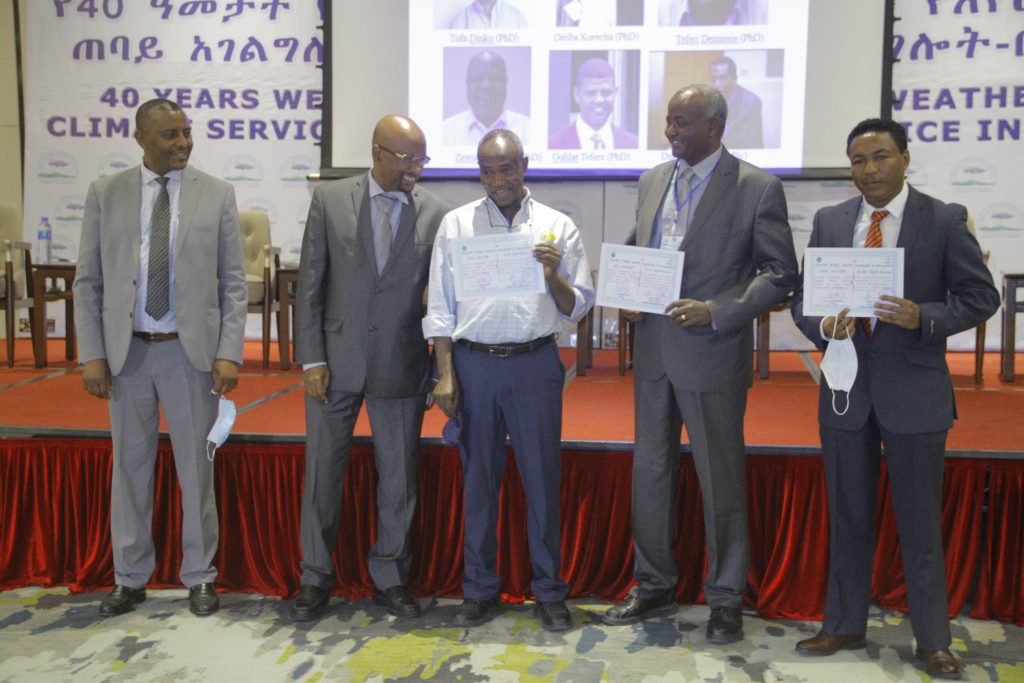 Tufa Dinku (middle) holds the document identifying IRI as a key partner in the development of Ethiopia’s National Framework for Climate Services, alongside several government officials on June 8, 2021. Five men stand in front of a red curtained stage that is set up for a panel with slides projected on a white screen. Three of the men hold white certificates, including Tufa Dinku, in the center.