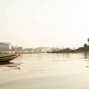 A Bangladesh man punts an empty boat across a river, with buildings of Dhaka in the background.