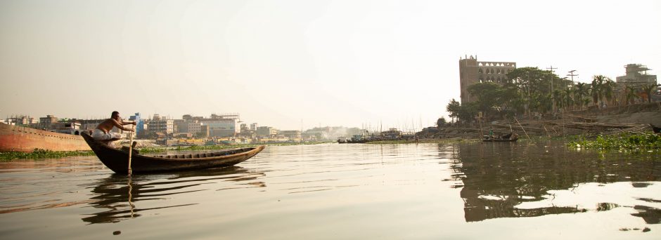 A Bangladesh man punts an empty boat across a river, with buildings of Dhaka in the background.