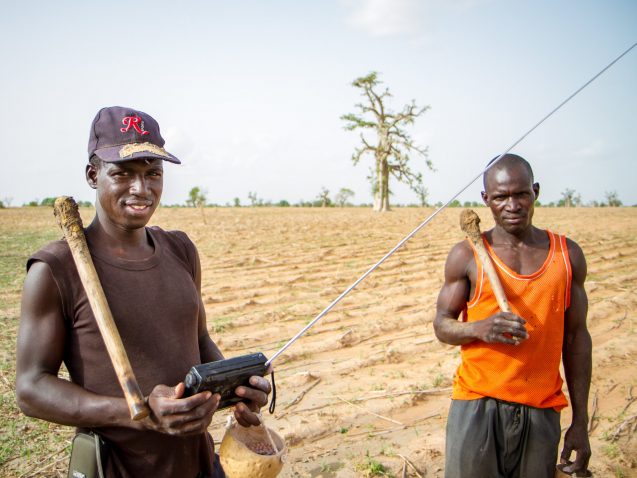 Two farmers in the village of Diouna, southern Mali, stand in a dry field. One holds a radio in his hands as they listen for weather bulletins.
