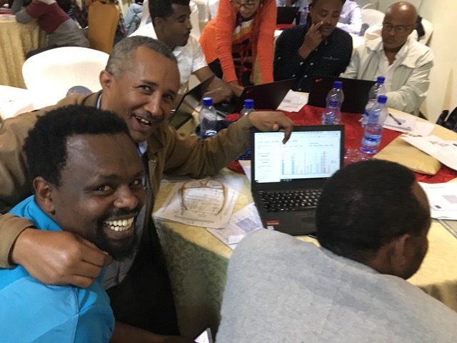 A group of Ethiopians sit around a round table with a tablecloth, papers and plastic water bottles. Two grinning men turn to face the camera, pointing to a laptop screen.