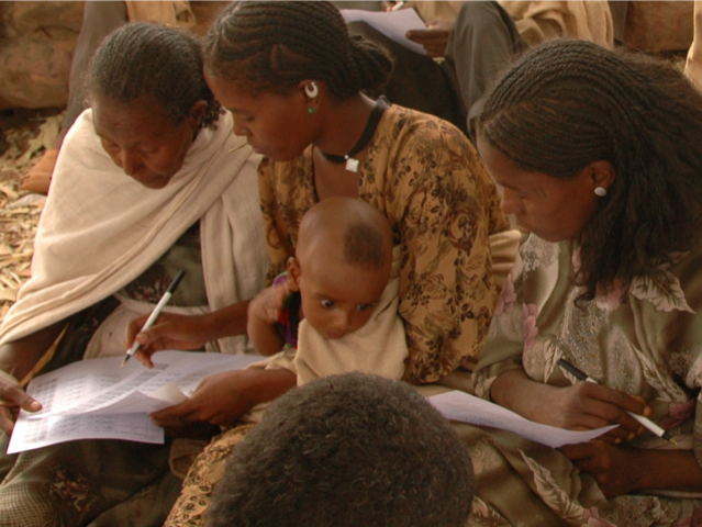 Three Ethiopian farmers look over worksheets at a focus group meeting. One of them holds a baby in her lap.